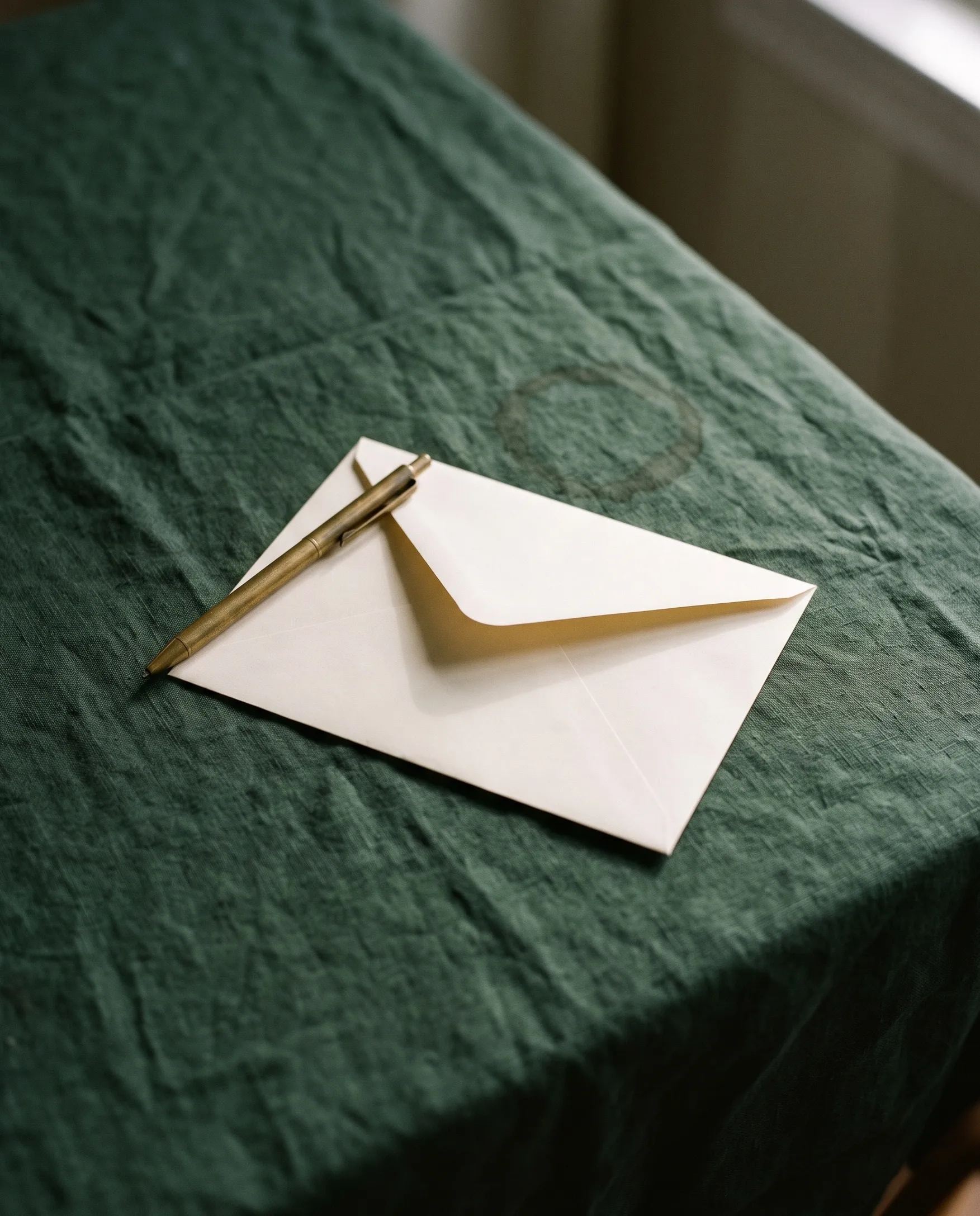 A cream envelope with its flap open and a brass pen lying across it on a crumpled forest-green linen tablecloth by a window