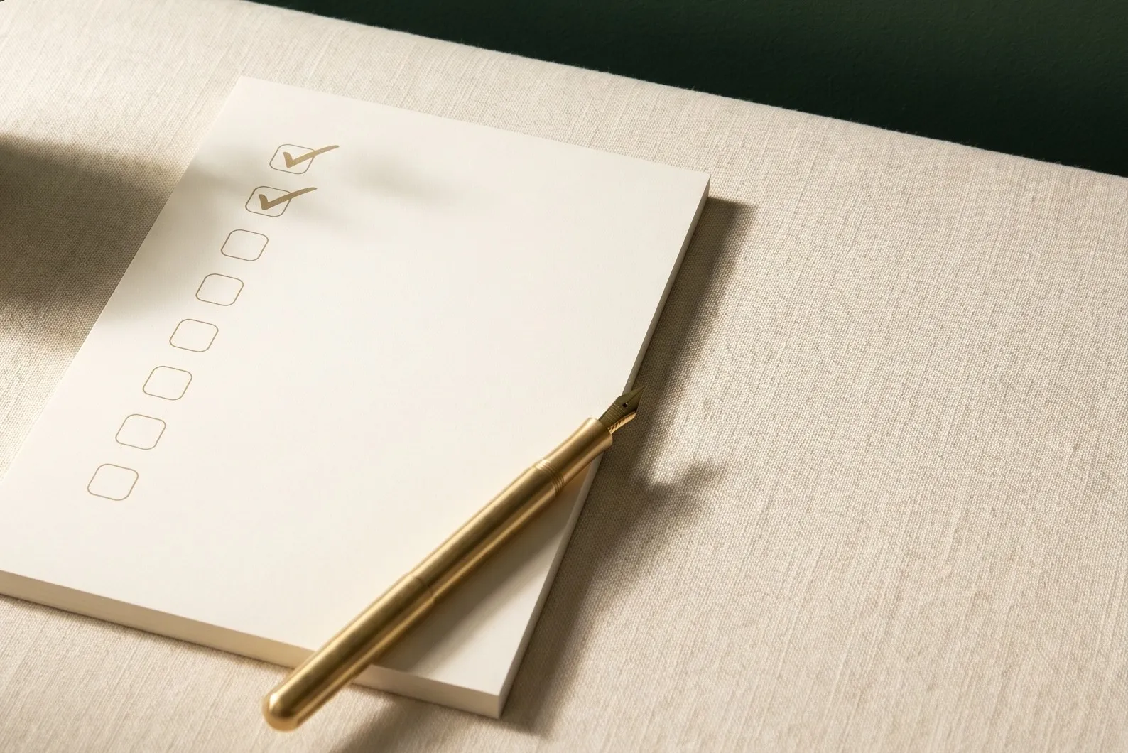 A cream notepad with a column of eight checkboxes, the top two ticked, and a brass fountain pen resting across its lower corner on a cream linen tablecloth, with a dark green wall behind