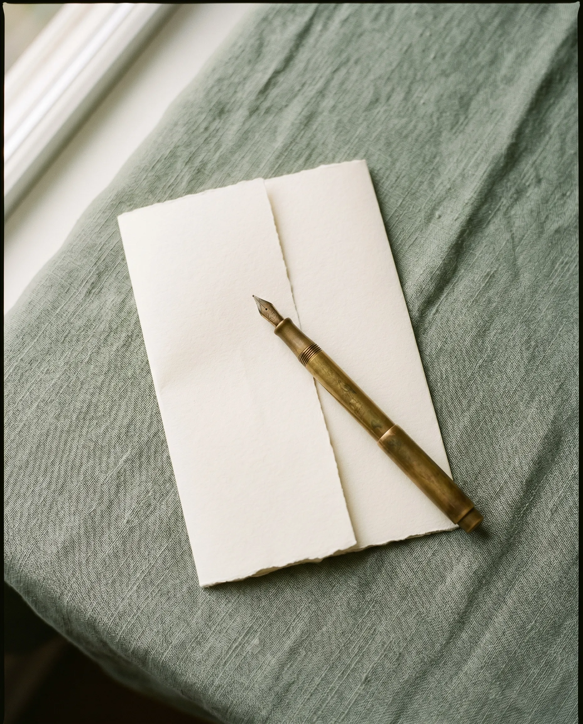 A folded sheet of cream deckle-edged paper with a brass fountain pen resting on it, laid on a sage-green linen cloth near a white window frame