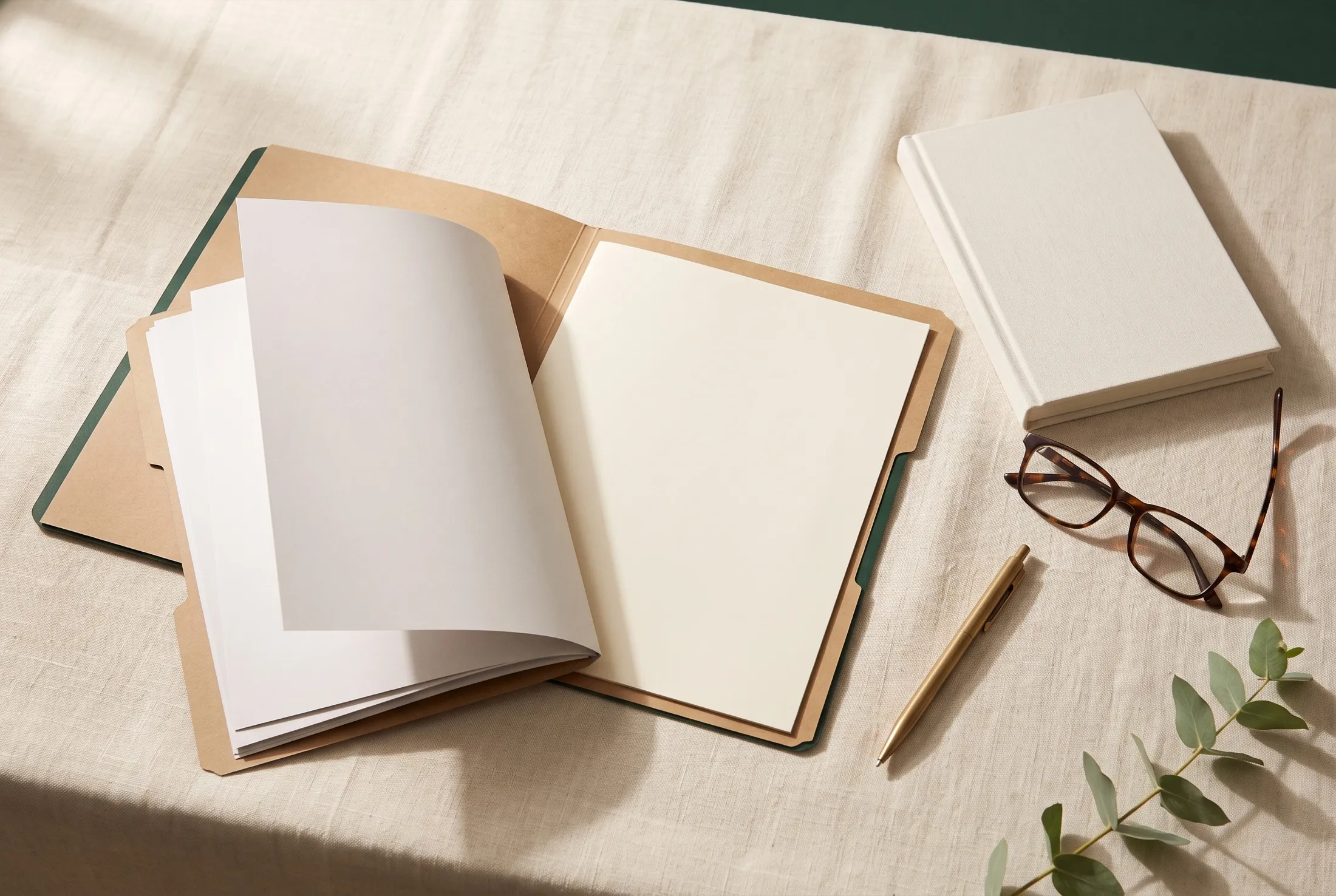 An open kraft document folder of blank pages beside a closed ivory notebook, tortoiseshell reading glasses and a brass pen on cream linen.